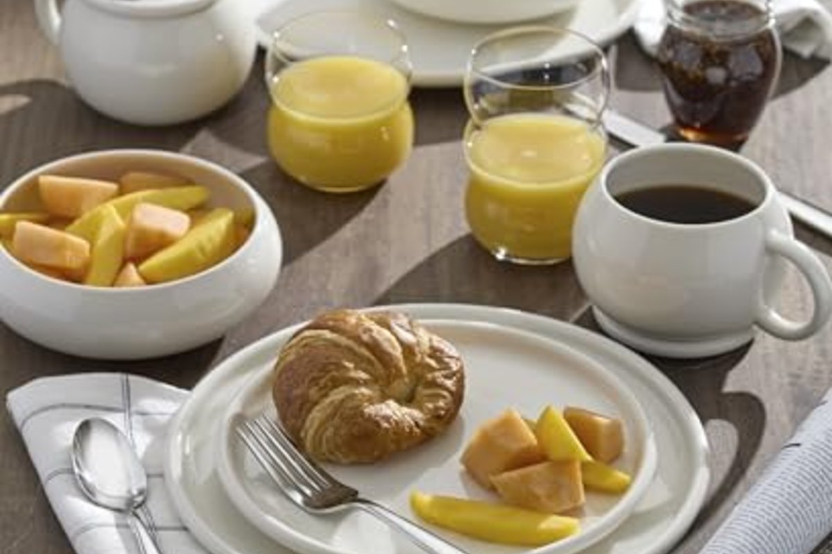 A breakfast table with a croissant, fruit, coffee and juice, cutlery rolled into a napkin